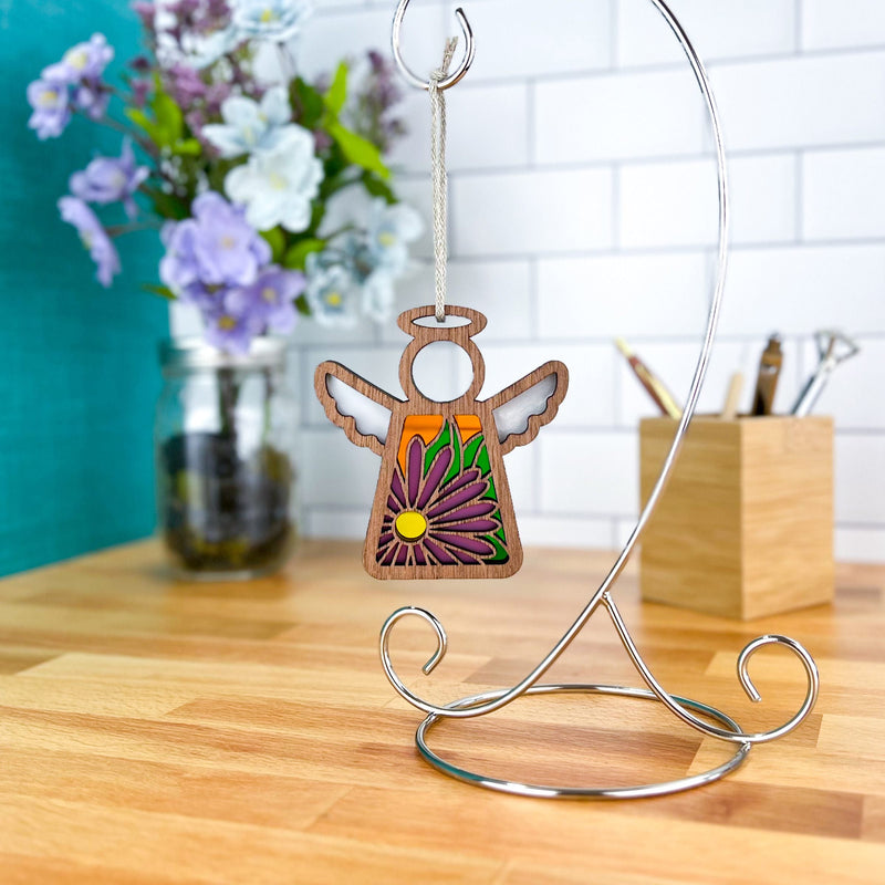 The Forged Flare® Aster Ornament, a 3.5" Mother's Angels® angel figurine featuring the September birth flower, hangs on a metal stand with a jar of flowers and wooden box on a countertop against white tile in the background.