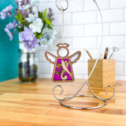 The Forged Flare® Honeysuckle Ornament | 3.5" (H) Angel Figurine | Mother's Angels® with white wings and a purple background hangs from a silver stand, beside a jar of honeysuckle flowers and art supplies on a wooden table.