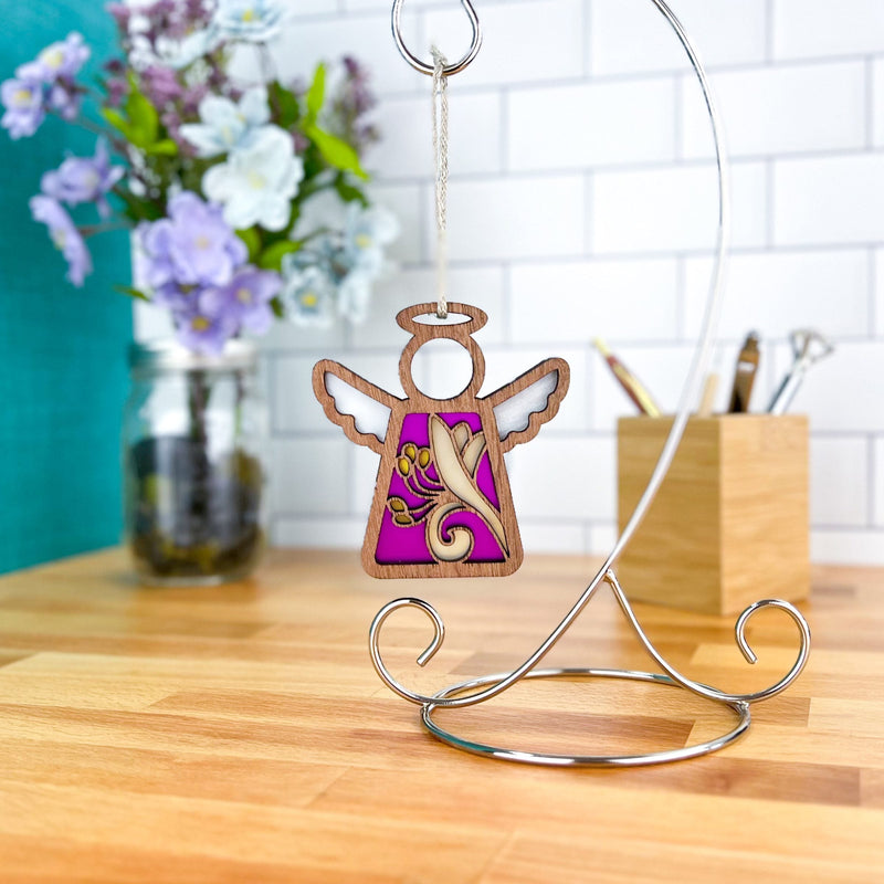The Forged Flare® Honeysuckle Ornament | 3.5" (H) Angel Figurine | Mother's Angels® with white wings and a purple background hangs from a silver stand, beside a jar of honeysuckle flowers and art supplies on a wooden table.