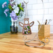 The Forged Flare® Hawthorn Ornament, a 3.5" angel figurine from Mother's Angels®, stands on a metal stand atop a wooden counter, with purple and white flowers and a box of pencils in the background.