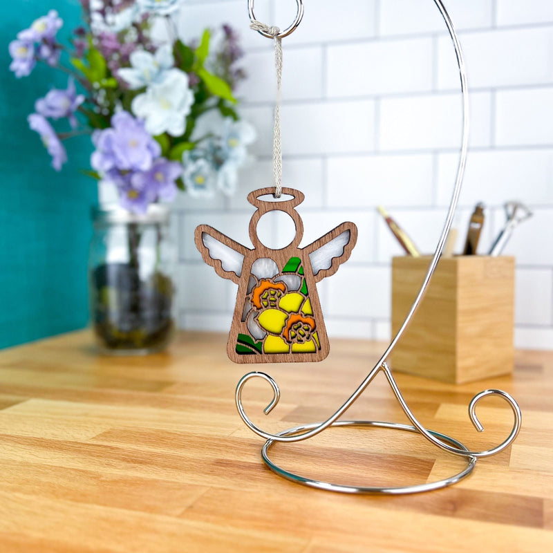 The Forged Flare® Daffodil Ornament | 3.5" (H) Angel Figurine from Mother's Angels® hangs from a silver stand on a wooden counter, with a mason jar of flowers and pen holder nearby—an ideal birth flower gift.