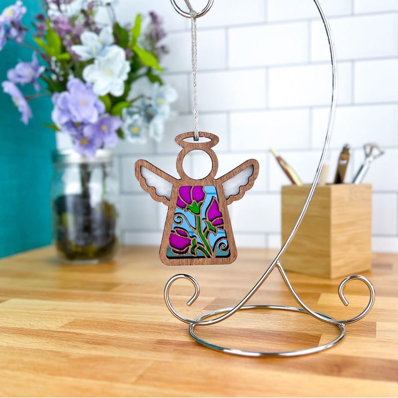 The Forged Flare® Sweet Pea Ornament—a 3.5" Mother's Angels® angel figurine with stained-glass detail—hangs on a metal stand atop a wooden desk, with sweet pea flowers and art supplies adding charm in the background.