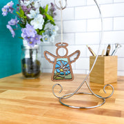 The Morning Glory Ornament, a 3.5" angel figurine from Forged Flare® Mother's Angels®, hangs from a metal stand on a wooden countertop. In the background are a jar of purple and white flowers and a wooden pencil holder.