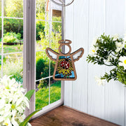 A Forged Flare® Mother's Angels® 3.5" angel figurine, the Ladybug and Dandelion Ornament, hangs by a window with white flowers on the sill and green trees and grass seen outside. Painted wings and a ladybug accent its body.