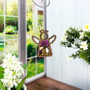 A Forged Flare® Mother's Angels® 3.5" Honey Bee Ornament angel figurine hangs indoors near a window, surrounded by white flowers and green plants, with a garden visible outside.