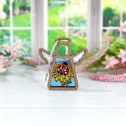 The Forged Flare® Mother's Angels® 3.5" Ladybug and Dandelion Ornament angel figurine stands on a white table, with blurred pink flowers and greenery near a window in the background.
