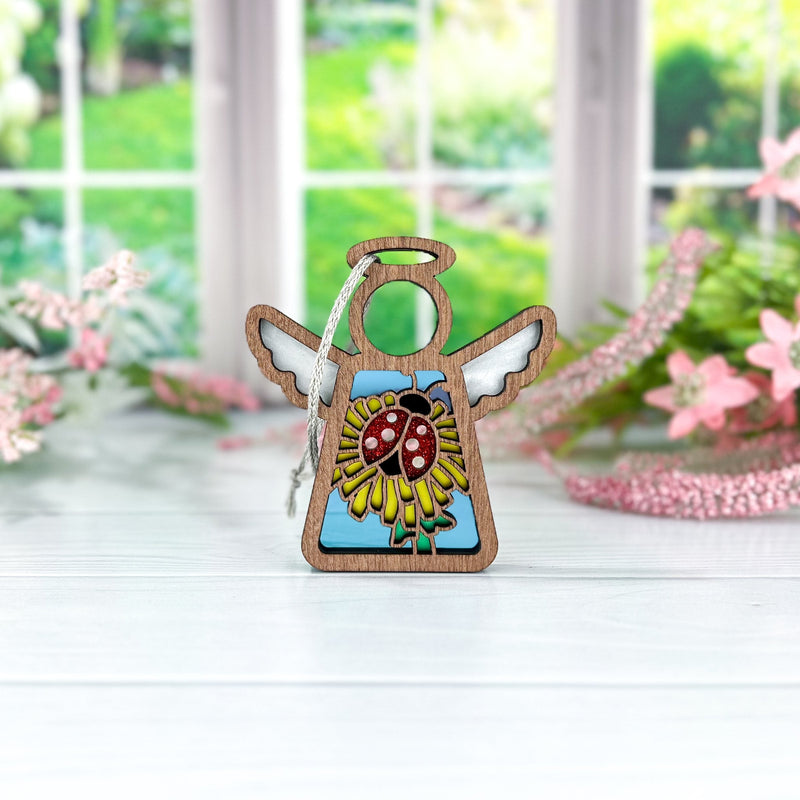 The Forged Flare® Mother's Angels® 3.5" Ladybug and Dandelion Ornament angel figurine stands on a white table, with blurred pink flowers and greenery near a window in the background.
