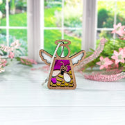 A 3.5" Mother's Angels® Honey Bee Ornament by Forged Flare®, featuring stained glass wings and a halo, stands on a white surface with pink flowers and greenery in the background—now available on Amazon.