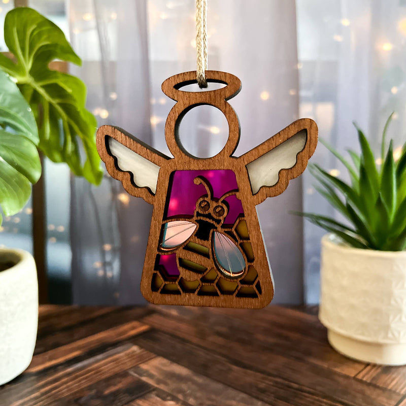 A Forged Flare® Warehouse Return - Honey Bee Ornament, a 3.5" Mother's Angels® angel figurine, hangs indoors by a string near green plants, glowing gently under lights as shown on Amazon.