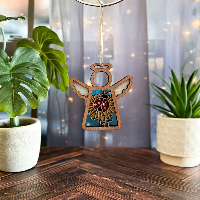 The Forged Flare® Ladybug and Dandelion Ornament is a 3.5" (H) Mother's Angels® figurine featuring an angel, perfect for cozy décor or as a Christmas tree ornament, displayed alongside plants and glowing string lights.