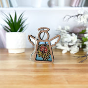 The Ladybug and Dandelion Ornament, a 3.5" Mother's Angels® figurine by Forged Flare®, features a wooden angel with ladybug and sunflower designs—a charming Christmas tree ornament with blurred flowers in the background.