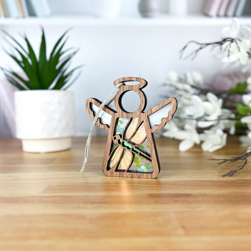 A Forged Flare® Warehouse Return - Dragonfly Ornament, a 3.5" Mother's Angels® stained glass angel figurine, stands on wood with plants, white flowers, and books in the background—reminiscent of an Amazon marketplace return.