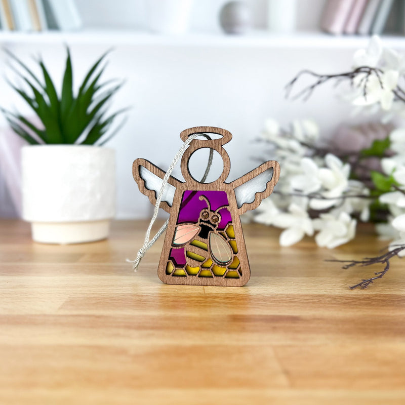 A Forged Flare® Warehouse Return - Honey Bee Ornament, a 3.5" Mother’s Angels® angel figurine with a stained glass bee design, stands on wood with a potted plant, white vase of flowers, and blurred books in the background.