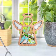 A Forged Flare® Mother's Angels® 3.5" dragonfly ornament hangs by a window with green plants and scissors in the background; features white wings and stained-glass details.