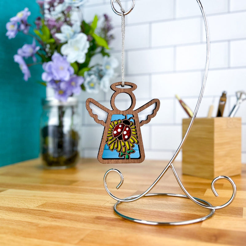The Forged Flare® Ladybug and Dandelion Ornament, a 3.5" Mother's Angels® wooden angel figurine, hangs on a metal stand near a vase of purple and white flowers and a wooden pen holder on the kitchen countertop.
