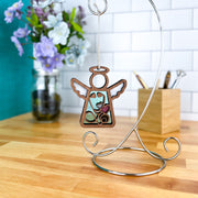 A Forged Flare® Mother's Angels® Doctor/Nurse Ornament with imperfections, a 3.5" wooden angel figurine, hangs on a silver stand atop a wood counter, with purple and white flowers and a branded collectible container holding pens nearby.