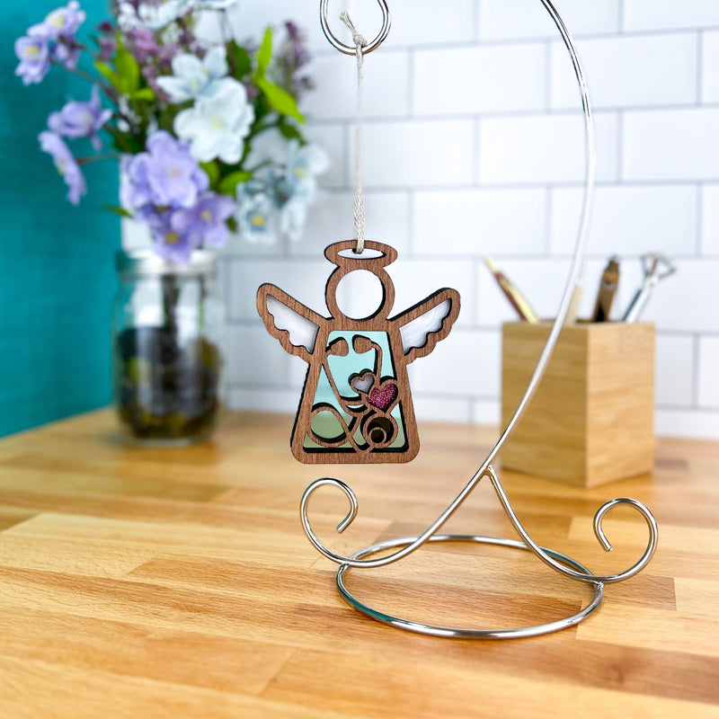 A Forged Flare® Mother's Angels® Doctor/Nurse Ornament with imperfections, a 3.5" wooden angel figurine, hangs on a silver stand atop a wood counter, with purple and white flowers and a branded collectible container holding pens nearby.