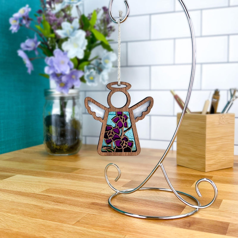 The Forged Flare® Larkspur Ornament, a 3.5" Mother's Angels® angel figurine with stained-glass-style wings and July's larkspur flowers, hangs from a silver stand beside a jar of purple and white blooms and a wooden utensil holder.