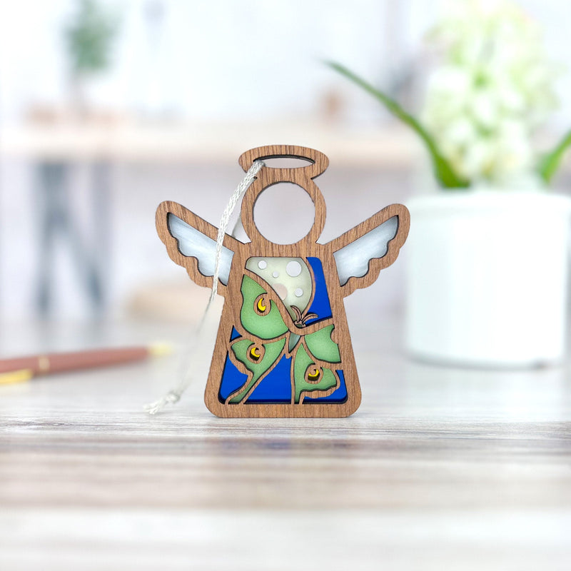 The Forged Flare® Luna Moth Ornament, a 3.5" Mother's Angels® wooden angel figurine, stands on a light wooden surface with softly blurred potted plant and pen in the background.