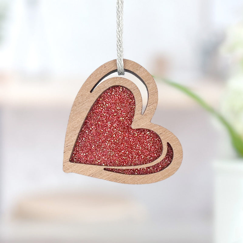 The Forged Flare® "I Love You" Red Heart Ornament, 2.5" (H), features a wooden heart with a red glittery center and sketch-style details, hanging by a white string against a softly blurred light background.