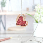The Forged Flare® "I Love You" Red Heart Ornament, 2.5"(H), hangs by a string, its glittery finish sparkling in the light. In the background, a white pot with green plants and a pen sit on a light-colored surface.