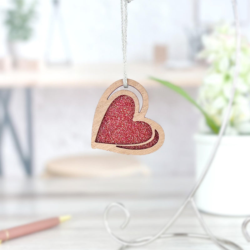 The Forged Flare® "I Love You" Red Heart Ornament, 2.5"(H), hangs by a string, its glittery finish sparkling in the light. In the background, a white pot with green plants and a pen sit on a light-colored surface.