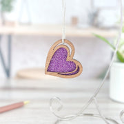 The Forged Flare® "I Love You" Purple Heart Ornament, 2.5" (H), features a sparkly center and hangs by a string, bringing charm to any desk with its enchanting look against a softly blurred backdrop of pencil and greenery in sketch style.