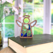 The Forged Flare® Luna Moth Ornament, a 3.5" Mother's Angels® angel figurine with stained-glass-style wings and dress, sits on a closed book by the window, with softly blurred pink flowers and greenery in the background.