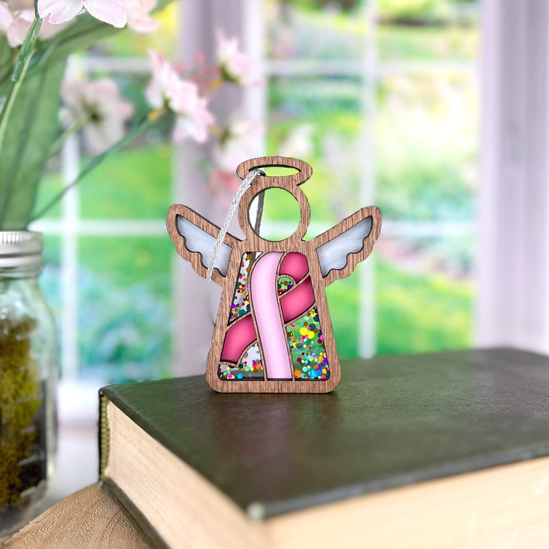 The Forged Flare® Mother's Angels® 3.5" Angel Figurine features a pink breast cancer awareness ribbon and colorful beads, making it a thoughtful charity ornament gift. Blurred flowers and greenery appear in the background by a window.