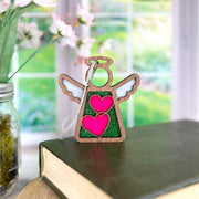 The Forged Flare® Limited Edition Interlocking Hearts Ornament - Series 8, a 3.5" Mother's Angels® figurine, stands on a closed book, with a glass jar of flowers and garden-view window softly blurred in the background.