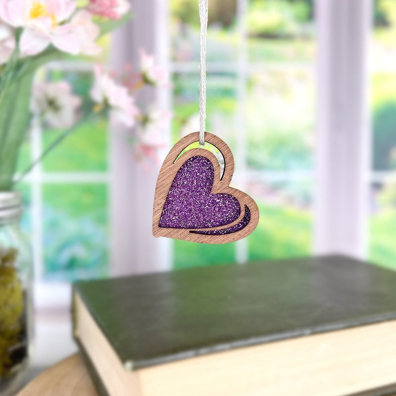 The Forged Flare® "I Love You" Purple Heart Ornament, 2.5" (H), with a glittery center, hangs in front of a window with blurred greenery and pink flowers; below sit a closed book and part of a glass jar for a serene touch.