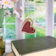 The Forged Flare® "I Love You" Red Heart Ornament, 2.5" (H), hangs in front of a blurred window beside a vase of flowers and closed books on a table.
