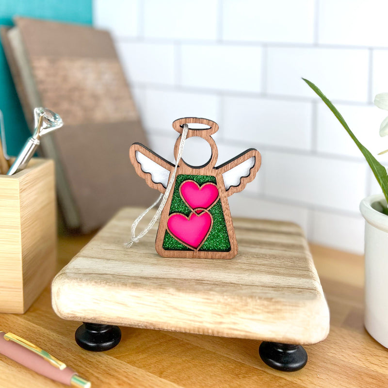 The Forged Flare® Limited Edition Interlocking Hearts Ornament - Series 8 is a 3.5" Mother's Angels® figurine with white wings, standing on a wooden platform amid books, a plant, and office supplies on a desk.