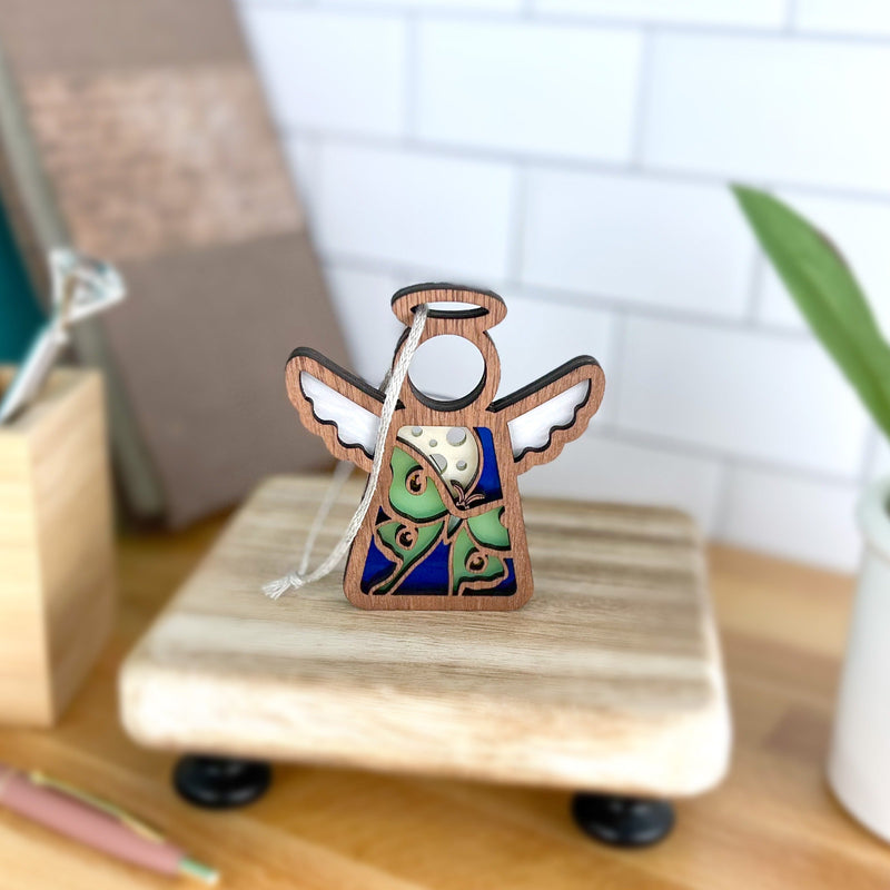 The Forged Flare® Luna Moth Ornament is a 3.5" Mother's Angels® figurine with white wings and a blue, brown, and green design. It stands on a wooden base and includes a hanging string, perfect as a Christmas tree ornament or decor piece.