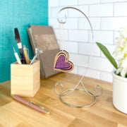 The "I Love You" Purple Heart Ornament (2.5" H) by Forged Flare® hangs on a metal stand atop a wooden desk, surrounded by art supplies, notebooks, a pen, and flowers in the background.