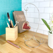 The Forged Flare® "I Love You" Red Heart Ornament, 2.5" (H), features a glittery red and gold sketch-style design and hangs from a silver stand on a wooden desk with art supplies, books, and flowers in the background.