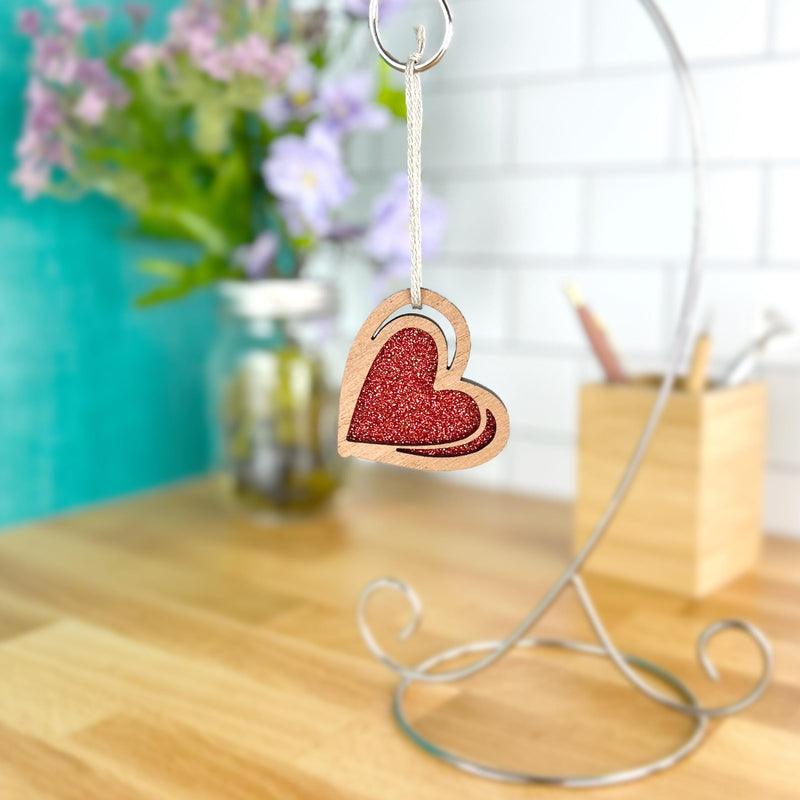 The Forged Flare® "I Love You" Red Heart Ornament, 2.5" (H), sparkles with red glitter as it hangs from a metal stand on a wooden desk. In the blurred background, flowers in a jar and a pencil container can be seen.