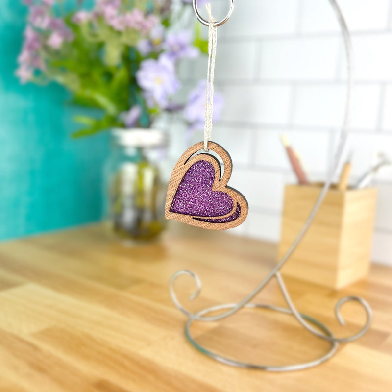 The Forged Flare® "I Love You" Purple Heart Ornament, 2.5" (H), features a glittery center and hangs on a stand. Behind it, a sketch-style scene shows flowers in a jar and pencils in a container on a wooden desk.