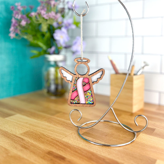 Pink ribbon angel ornament hangs on a stand, a symbol of hope for breast cancer awareness, amidst a bright setting.