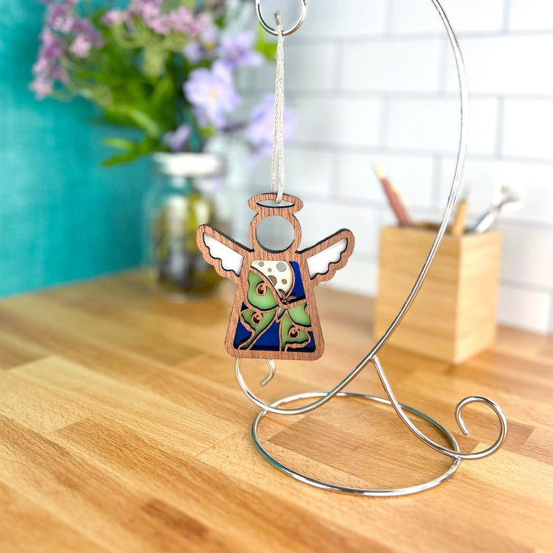 The Luna Moth Ornament | 3.5" (H) Angel Figurine by Forged Flare® from the Mother's Angels® collection hangs from a silver stand on a wooden countertop, with flowers in a jar and a pencil-filled wooden container in the background.