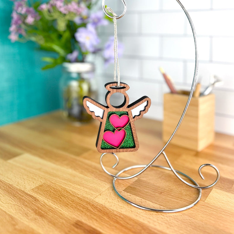 The Limited Edition Interlocking Hearts Ornament – Series 8 | 3.5" Angel Figurine by Forged Flare® hangs from a silver stand on a wooden countertop, with flowers and a tiled wall highlighting this charming Mother's Angels® display.