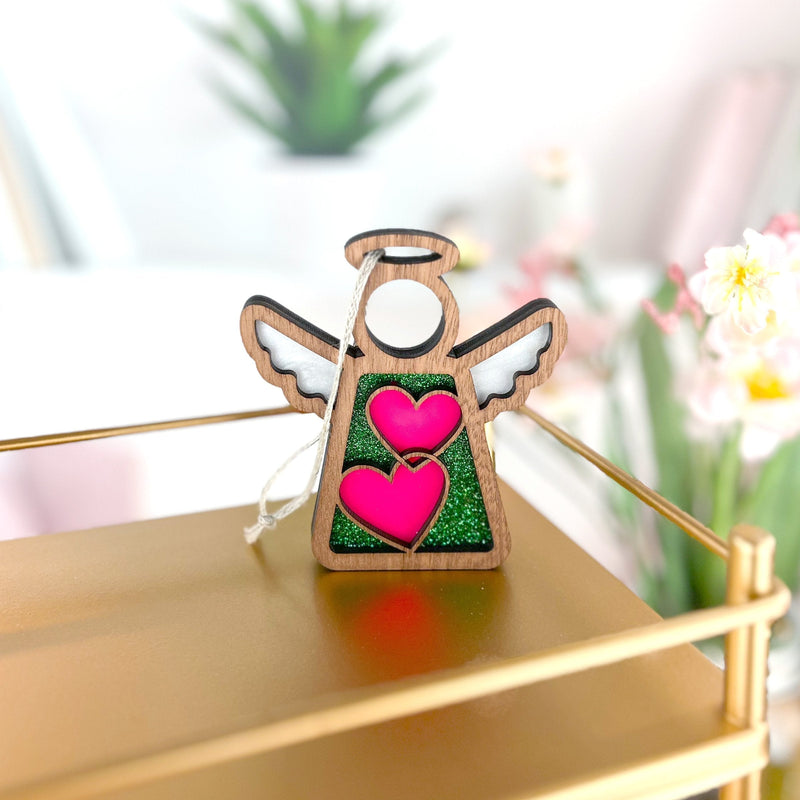 The Forged Flare® Limited Edition Interlocking Hearts Ornament - Series 8 is a 3.5" Mother's Angels® figurine with white wings, pink hearts, and green glitter, shown on a gold surface with flowers and a potted plant blurred in the background.