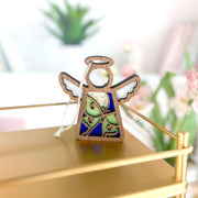 The Luna Moth Ornament, a 3.5" angel figurine by Forged Flare® Mother's Angels®, features blue and green details and hangs by a white string on a gold shelf with blurred plants and flowers in the background.