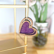 The Forged Flare® "I Love You" Purple Heart Ornament, 2.5" (H), hangs from a string on a gold metal shelf, its glittery center shining with a blurred jar of flowers in the background.