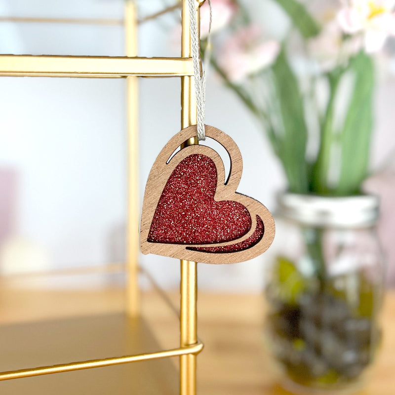 The "I Love You" Red Heart Ornament, 2.5" (H) by Forged Flare® hangs from a gold metal shelf; its glittery center shines as blurred flowers in a jar appear in the background.