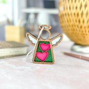 The Forged Flare® Limited Edition Interlocking Hearts Ornament - Series 8 is a 3.5" wooden Mother's Angels® figurine with white wings, green glitter center, and pink interlocking hearts, perfect as a festive Christmas tree ornament.