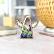 The Forged Flare® Luna Moth Ornament is a 3.5" Mother's Angels® wooden angel figurine with white wings and a colorful abstract design, displayed on a light desk alongside art supplies and a woven lamp.