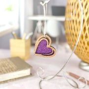 The Forged Flare® "I Love You" Purple Heart Ornament, 2.5" (H), features a wooden heart with a sparkling purple glitter center on a string. Behind it, artistic sketches of a desk, pencils, notepad, and lamp create creative ambiance.