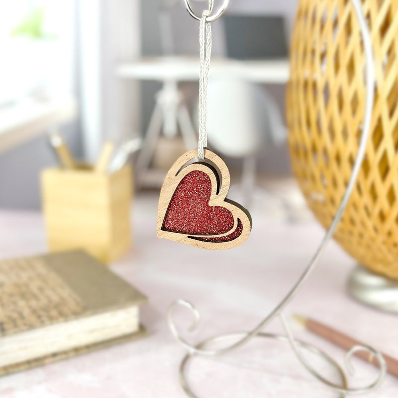 The Forged Flare® "I Love You" Red Heart Ornament, 2.5" (H), featuring a glittery center and sketch-style design, hangs from a stand; behind it, blurred desk items and warm lighting create a cozy workspace vibe.