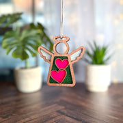 The Forged Flare® Limited Edition Interlocking Hearts Ornament – Series 8 features a 3.5" wooden angel figurine with green glitter, hanging by a string. Blurred potted plants and soft lights create a cozy backdrop on a wooden surface.